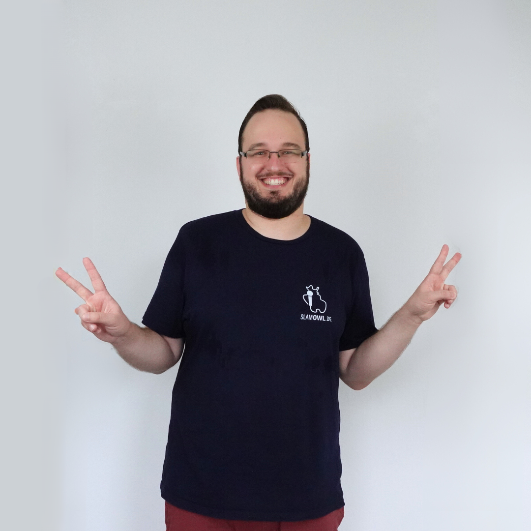 Ein Mann mit Brille und Bart lächelt und hält beide Hände zum Peace-Zeichen hoch, während er ein marineblaues T-Shirt mit dem kleinen Slam OWL-Logo aus der „T-Shirt Poetry Slam – Slam OWL – Logo & Eule“-Kollektion von Artbookings/Shirtigo trägt.