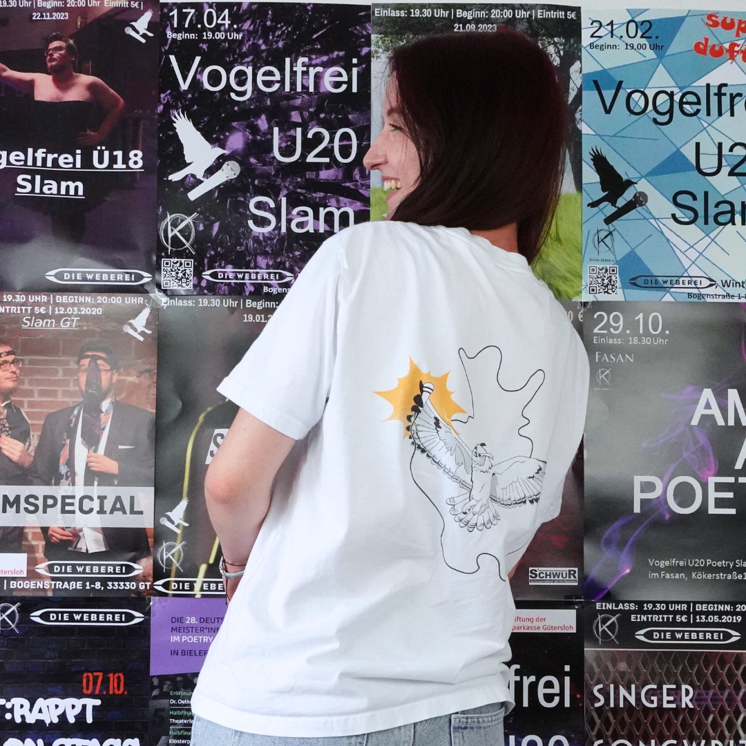 Eine Person, die ein „T-Shirt Poetry Slam – Slam OWL – Logo & Eule – schwarz auf weiß | Bio-Qualität“ von Artbookings/Shirtigo trägt, steht leicht zur Seite gedreht vor einer Wand, die mit Veranstaltungsplakaten bedeckt ist. Darunter befindet sich ein auffälliger Flyer zum kommenden Poetry Slam OWL in Ostwestfalen-Lippe.