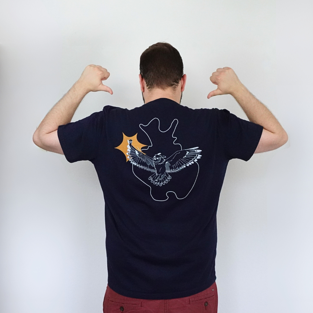 Eine Person mit kurzen Haaren trägt ein dunkles T-Shirt Poetry Slam - Slam OWL von Artbookings/Shirtigo und präsentiert stolz die weiß-orangefarbene Illustration eines Adlers und Sterns auf dem Bio-Qualitätsshirt. Sie zeigt selbstbewusst mit dem Daumen nach hinten auf das Design und betont stolz ihre ostwestfalen-lippische Herkunft in einem Stil, der perfekt zu einem Poetry Slam passt.