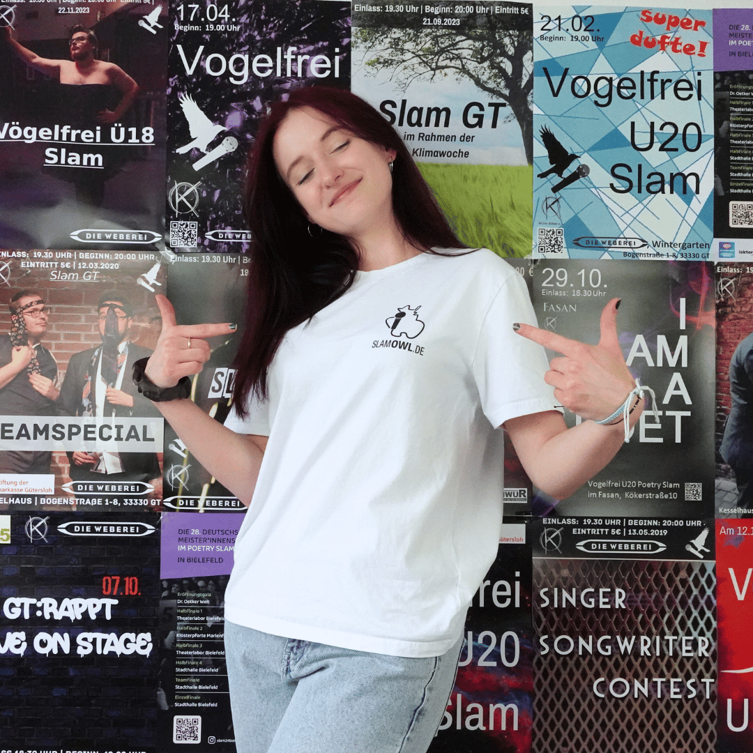 Eine Person mit einem T-Shirt Poetry Slam – Slam OWL – Logo & Eule – schwarz auf weiß von Artbookings/Shirtigo posiert mit erhobenem Daumen vor einer Wand, die mit verschiedenen bunten Veranstaltungsplakaten bedeckt ist, unter anderem für den Poetry Slam OWL in Ostwestfalen-Lippe.