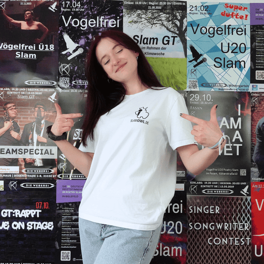 Eine Person mit einem T-Shirt Poetry Slam – Slam OWL – Logo & Eule – schwarz auf weiß von Artbookings/Shirtigo posiert mit erhobenem Daumen vor einer Wand, die mit verschiedenen bunten Veranstaltungsplakaten bedeckt ist, unter anderem für den Poetry Slam OWL in Ostwestfalen-Lippe.