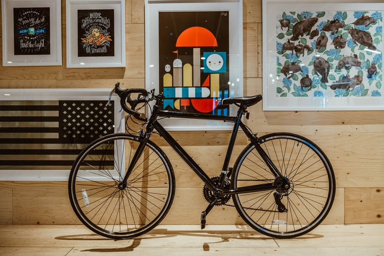 Ein schwarzes Rennrad ist drinnen an einer Holzwand geparkt, die mit verschiedenen gerahmten Kunstwerken und Postern dekoriert ist.