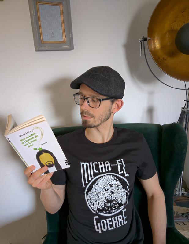 Ein Mann mit Brille und Mütze sitzt auf einem Stuhl und liest ein Buch. Er trägt ein schwarzes T-Shirt mit einer Grafik, auf der "MICHA-EL GOEHRE" steht. Im Hintergrund ist eine große Lampe zu sehen.