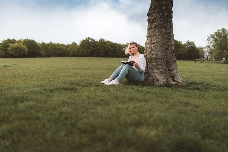 Eine Person sitzt im Gras, lehnt sich an einen Baum und liest ein Buch in einem weitläufigen, grünen Park unter einem teilweise bewölkten Himmel.