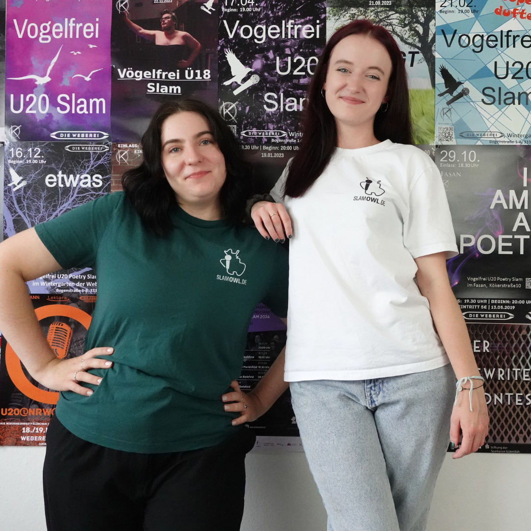 Zwei Frauen stehen vor einer Wand voller Plakate, eine trägt das T-Shirt Poetry Slam - SLAM OWL - Logo & Eule in grün mit schwarzer Hose, die andere trägt die weiße Version des gleichen T-Shirts mit passender weißer Hose. Beide lächeln, die Hände in die Hüften gestemmt, und genießen sichtlich den Moment vor dem Poetry Slam. Die T-Shirts im Bioqualitätsdesign von Artbookings/Shirtigo verleihen ihrem Outfit eine stilvolle Note.