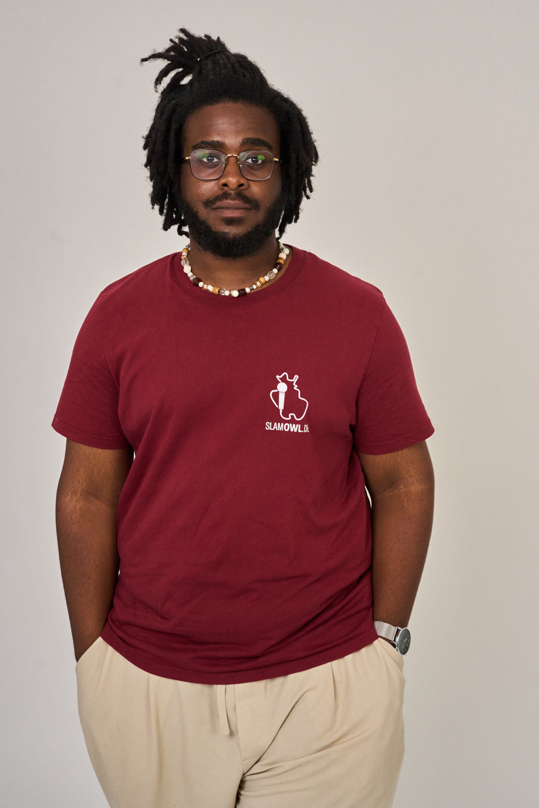 Ein Mann mit kurzen Dreadlocks, Brille und einer Perlenkette trägt das kastanienbraune "T-Shirt Poetry Slam - SLAM OWL - Logo & Eule - weiß auf Farbe" von Artbookings/Shirtigo mit beiger Hose vor einem schlichten Hintergrund.