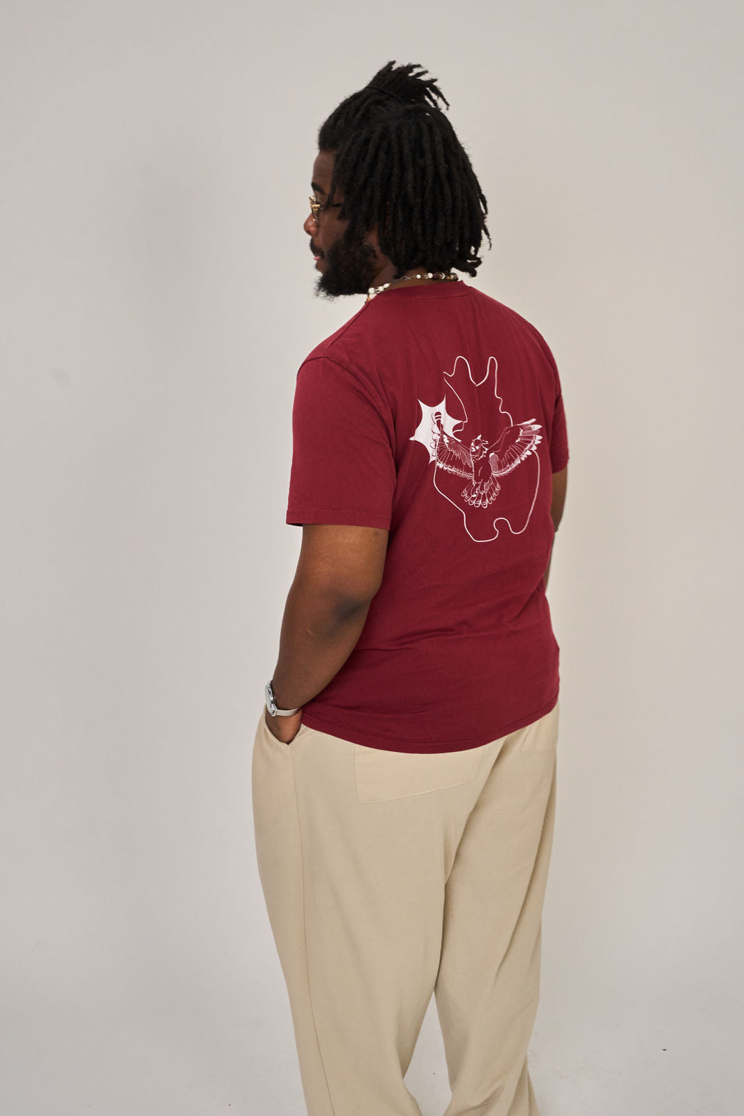 Eine Person steht mit dem Rücken zur Kamera vor einem schlichten, hellen Hintergrund. Sie trägt eine beigefarbene Hose und das kastanienbraune Artbookings/Shirtigo "T-Shirt Poetry Slam - SLAM OWL" in Bioqualität mit einem weißen Eulenlogo auf der Rückseite.