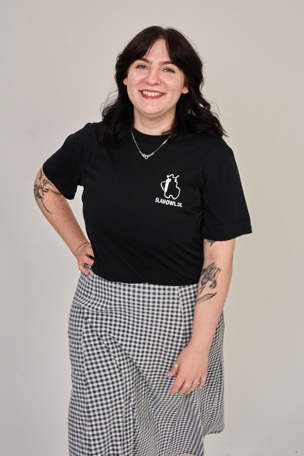 Eine Person mit dunklen Haaren trägt das Artbookings/Shirtigo T-Shirt Poetry Slam - SLAM OWL mit einem weißen Eulenlogo auf Schwarz, gepaart mit einem schwarz-weiß karierten Rock, vor einem unifarbenen Hintergrund.
