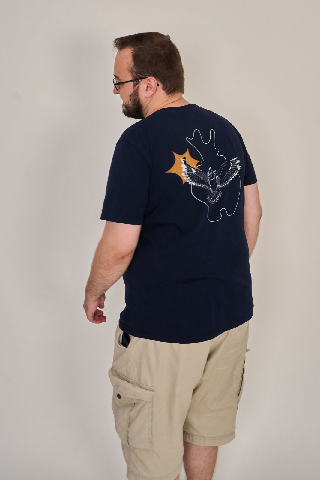 Ein Mann, der ein marineblaues Artbookings/Shirtigo-T-Shirt Poetry Slam - SLAM OWL mit einem weißen Eulenlogo und einer geplatzten Grafik auf dem Rücken sowie eine helle Cargo-Shorts trägt, steht mit dem Rücken zur Kamera.