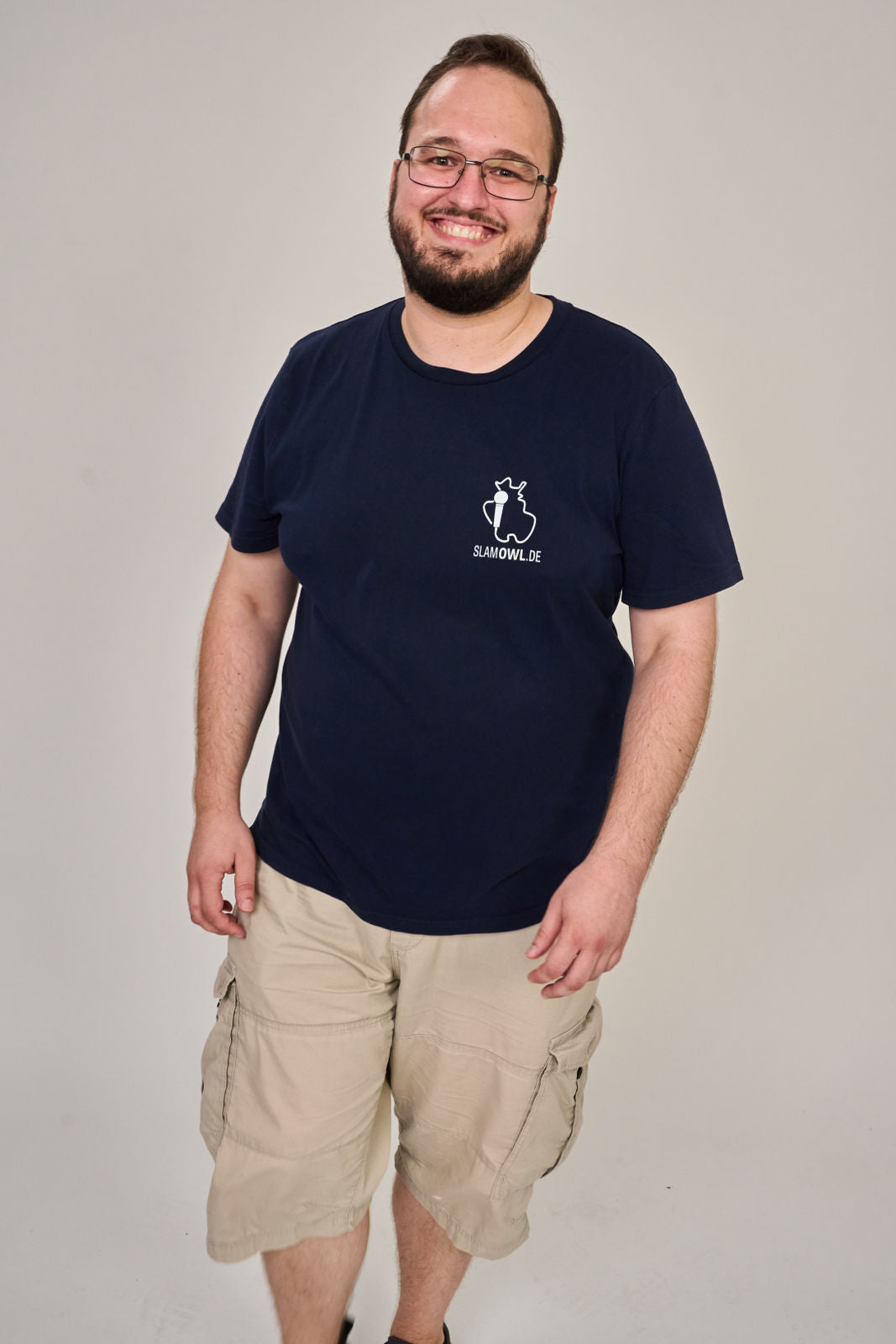 Ein Mann mit Brille, Bart und kurzen Haaren lächelt vor einem unifarbenen Hintergrund. Er trägt ein marineblaues Artbookings/Shirtigo T-Shirt Poetry Slam - SLAM OWL mit weißem Logo und Eule-Motiv, gepaart mit hellen Cargo-Shorts.