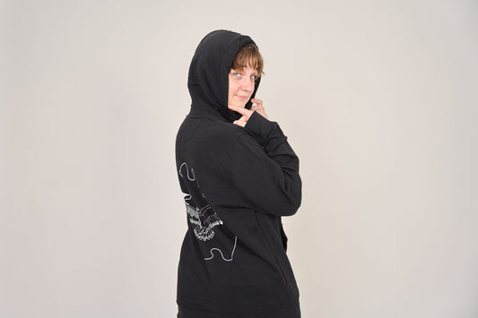 Eine Person posiert vor einem schlichten Hintergrund, trägt den Artbookings/Shirtigo Hoodie Poetry Slam - SLAM OWL in Bio-Qualität, mit weißem Eulen-Logoaufdruck auf dem Rücken, die Kapuze hochgezogen und schaut über die Schulter.
