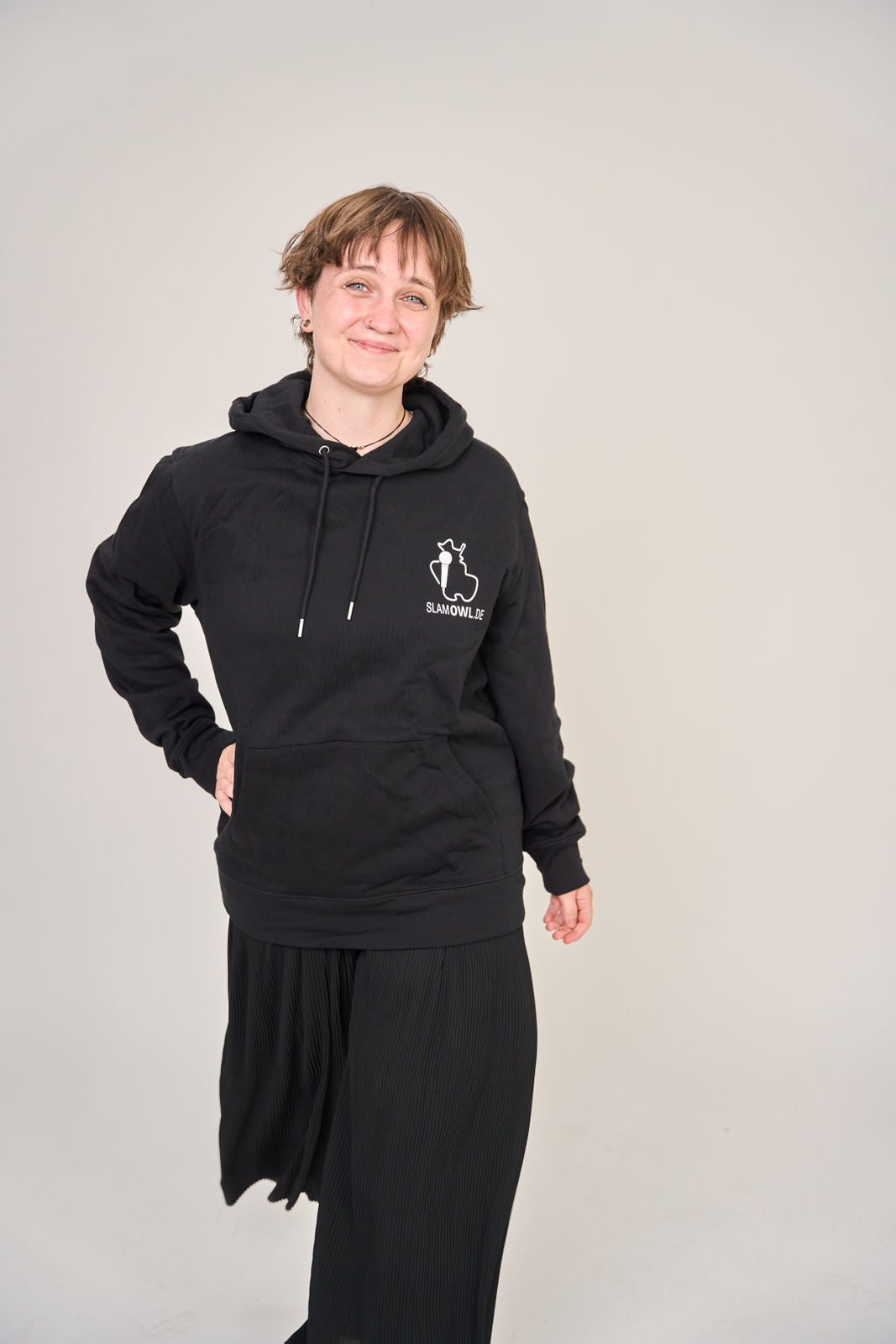 Eine Person steht vor einem schlichten Hintergrund, trägt den Hoodie Poetry Slam - SLAM OWL mit weißem Aufdruck von Artbookings/Shirtigo (Bio-Qualität) und schwarzer Hose und lächelt leicht in die Kamera.