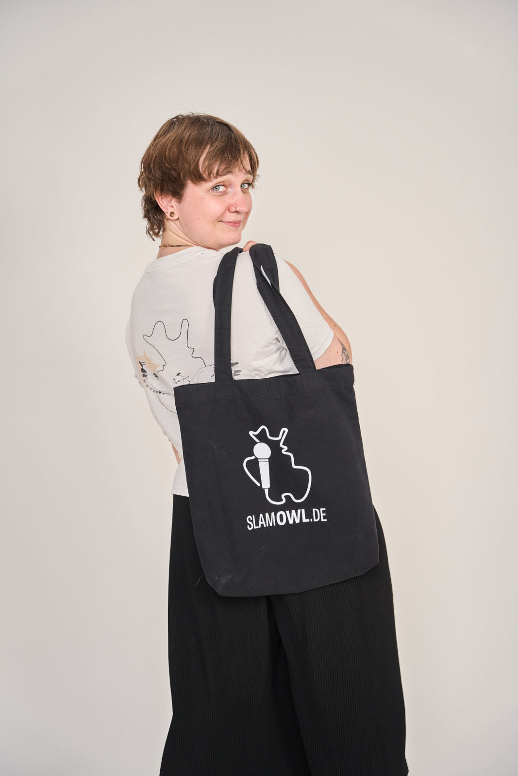 Eine Person mit kurzen Haaren schaut über ihre Schulter und trägt die Artbookings/Shirtigo Baumwoll-Tragetasche Slam OWL mit Logo & Eule - dark, eine schwarze Tragetasche mit weißer Eulengrafik und Logo für den Poetry Slam Ostwestfalen-Lippe.