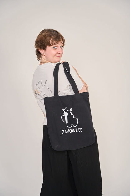 Eine Person mit kurzen Haaren schaut über ihre Schulter und trägt die Artbookings/Shirtigo Baumwoll-Tragetasche Slam OWL mit Logo & Eule - dark, eine schwarze Tragetasche mit weißer Eulengrafik und Logo für den Poetry Slam Ostwestfalen-Lippe.