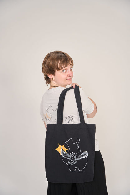 Eine Person mit kurzen Haaren posiert mit der schwarzen Baumwoll-Tragetasche Slam OWL mit Logo & Eule von Artbookings/Shirtigo. Ihr helles T-Shirt passt zum Design des Poetry Slam Ostwestfalen-Lippe und zelebriert den Slam OWL-Stil.