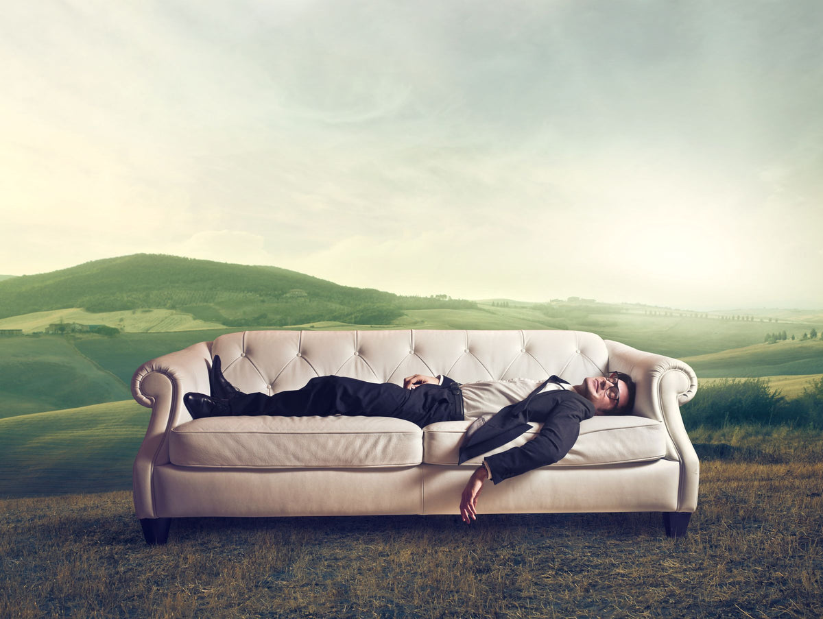 Mann im dunklen Anzug liegt auf einem hellen Sofa mitten auf einer grünen Hügelwiese