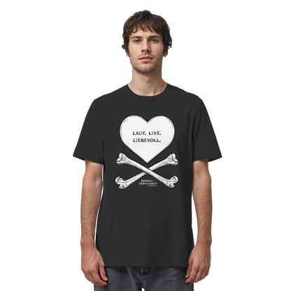 Ein Mann trägt das „Laut. Live. Liebevoll. – T-Shirt mit Statement | Weiß auf Schwarz“ von Artbookings/Shirtigo – ein schwarzes Shirt mit einem weißen Herz über gekreuzten Knochen, in dessen Inneren die Worte „Laut. Live. Liebevoll.“ stehen und das den kühnen Geist von „Krawall + Zärtlichkeit“ verkörpert – ideal für Fans des Poetry Slam Essen.