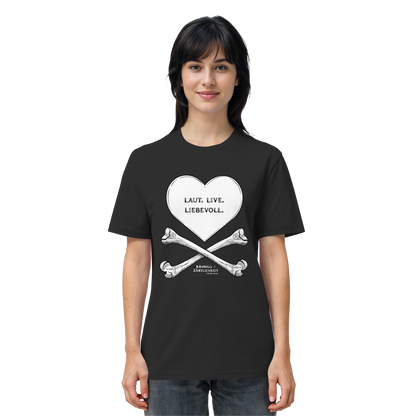 Eine Frau blickt mit einem leichten Lächeln nach vorne und trägt das Artbookings/Shirtigo-Statement-T-Shirt „Laut. Live. Liebevoll.“ (Weiß auf Schwarz). Das schwarze Shirt zeigt eine auffällige Grafik aus einem Herz und gekreuzten Knochen mit der Aufschrift „LAUT. LIEB. LIEBEVOLL.“ auf Deutsch – ein Motiv, das den Geist von „Krawall + Zärtlichkeit“ verkörpert, der bei Veranstaltungen wie dem Poetry Slam Essen beliebt ist.