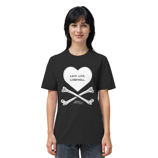 Eine Frau blickt mit einem leichten Lächeln nach vorne und trägt das Artbookings/Shirtigo-Statement-T-Shirt „Laut. Live. Liebevoll.“ (Weiß auf Schwarz). Das schwarze Shirt zeigt eine auffällige Grafik aus einem Herz und gekreuzten Knochen mit der Aufschrift „LAUT. LIEB. LIEBEVOLL.“ auf Deutsch – ein Motiv, das den Geist von „Krawall + Zärtlichkeit“ verkörpert, der bei Veranstaltungen wie dem Poetry Slam Essen beliebt ist.