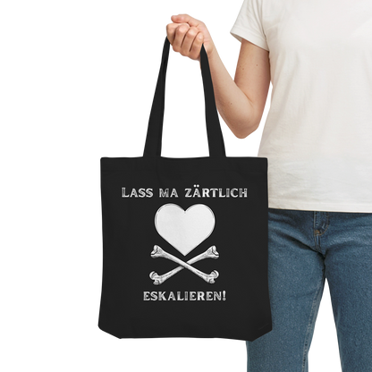 Person mit der schwarzen „Krawall + Zärtlichkeit Poetry Slam – Baumwolltragetasche – Lass ma zärtlich eskalieren“ von Artbookings/Shirtigo – weißes Herz über Knochen, perfekt für Poetry Slams.